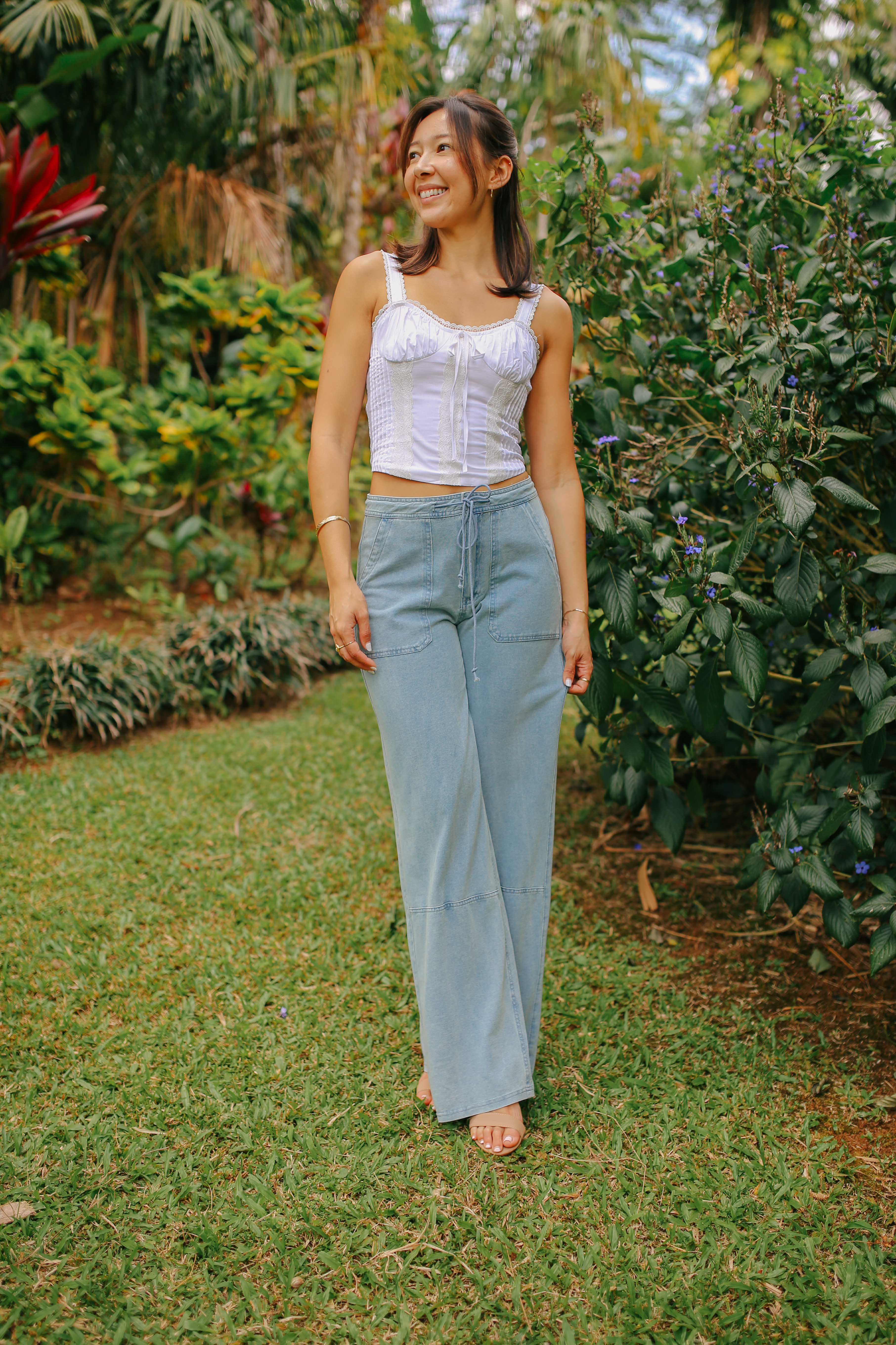 A smiling young woman standing in a lush garden. She is wearing a white sleeveless top with lace detailing and light blue, wide-leg denim pants. Her hair is half-up and she is looking to the side.