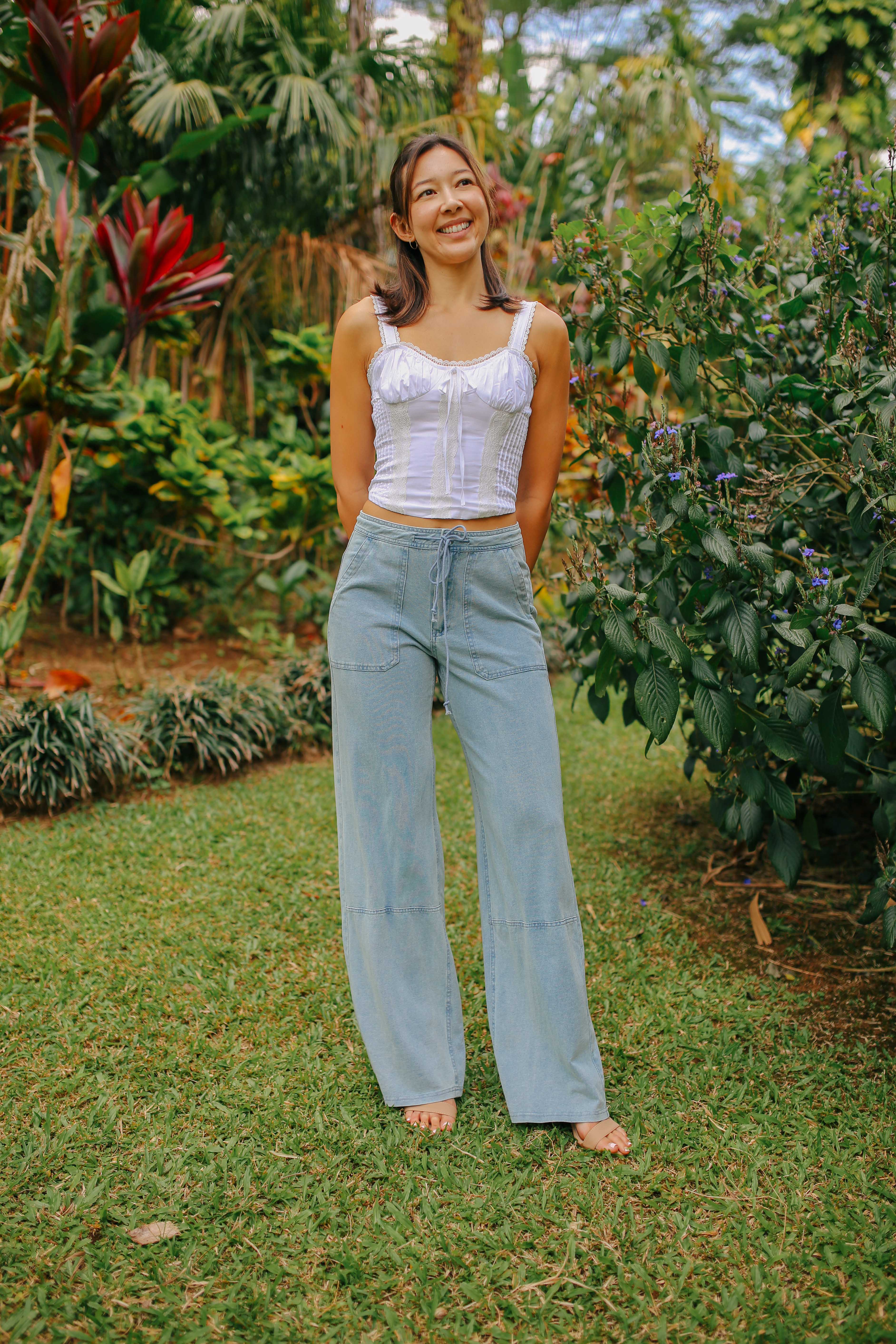 A woman standing on grass wearing a white tank top and light blue drawstring cargo pants with a wide leg.