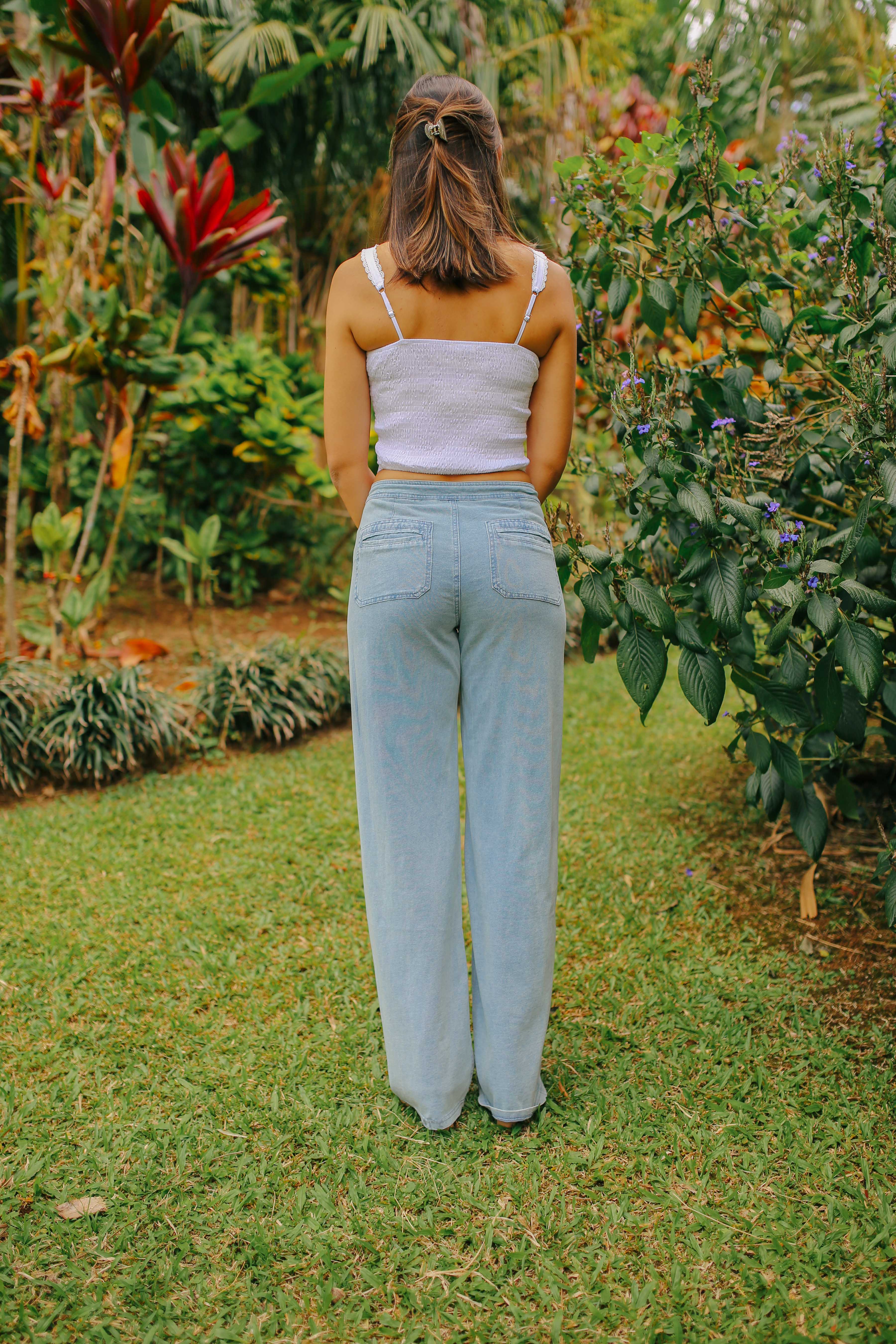 A full-length, back-facing shot of a  woman in a garden, wearing a white lace tank top and light blue, wide-leg denim pants.