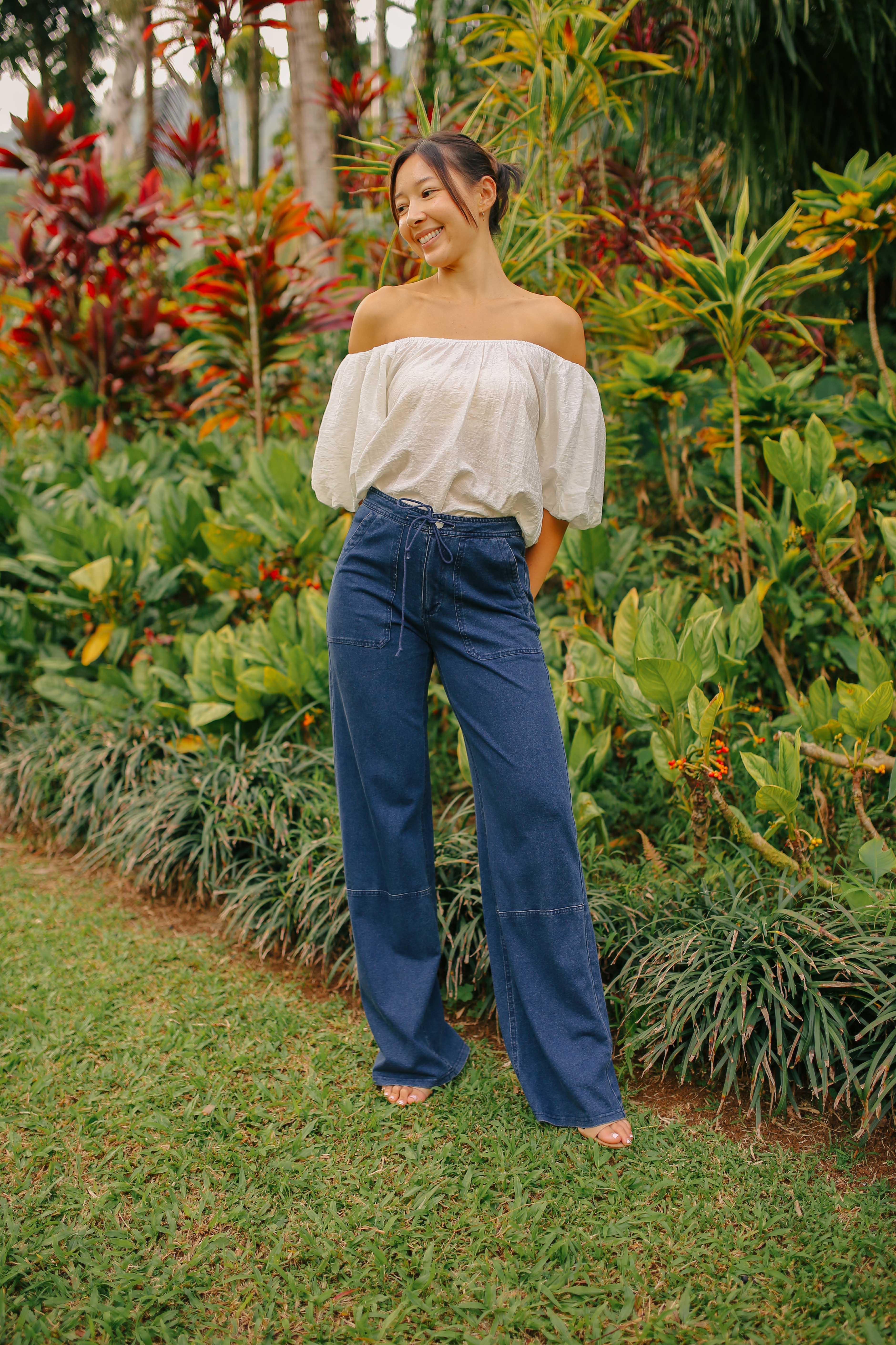 A woman standing outdoors wearing blue wide-leg cargo pants and a white off-shoulder top.