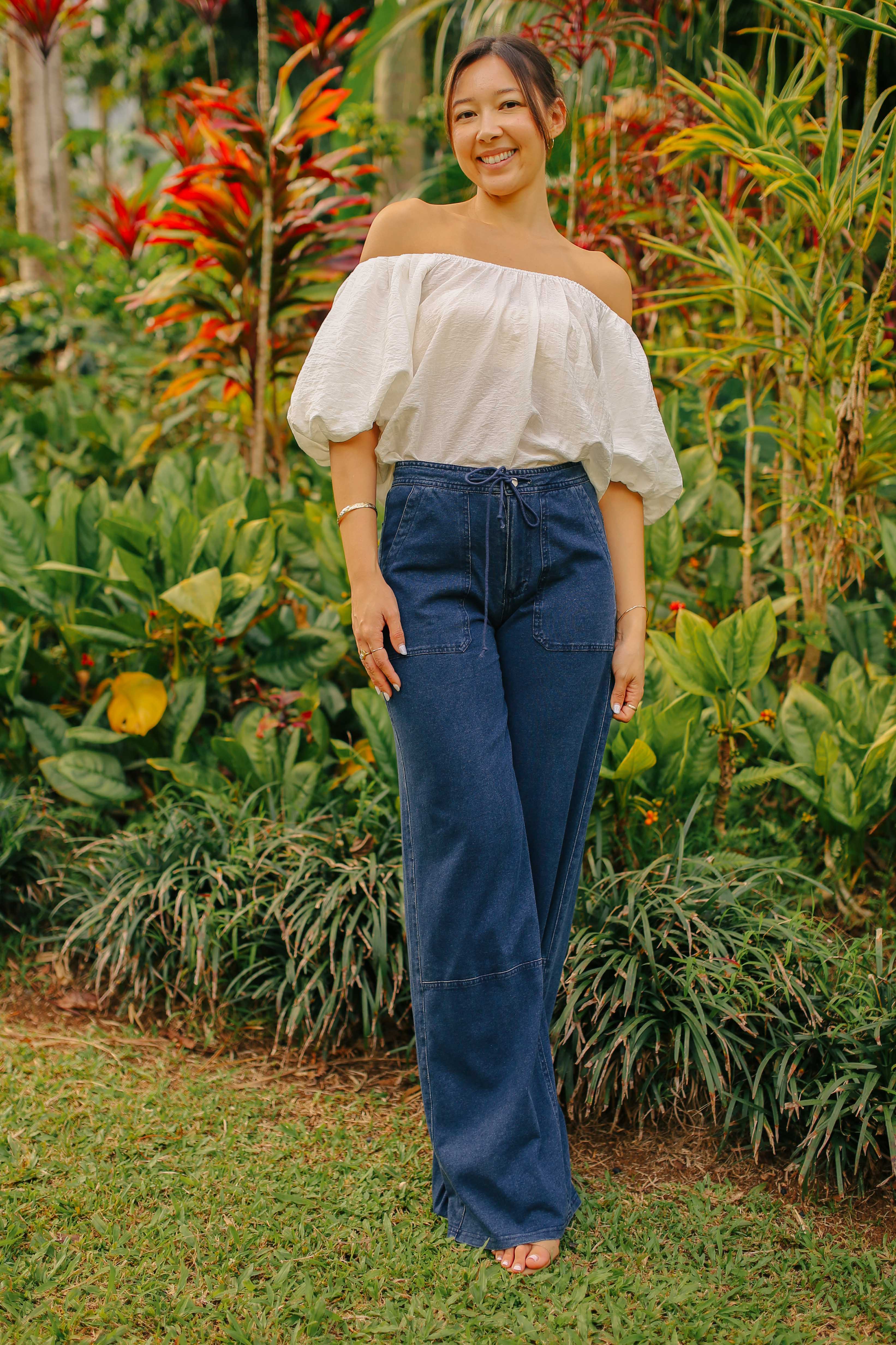 A young woman standing in a lush, green garden. She is wearing a white off-the-shoulder, short-sleeved top and dark blue, wide-leg denim pants.