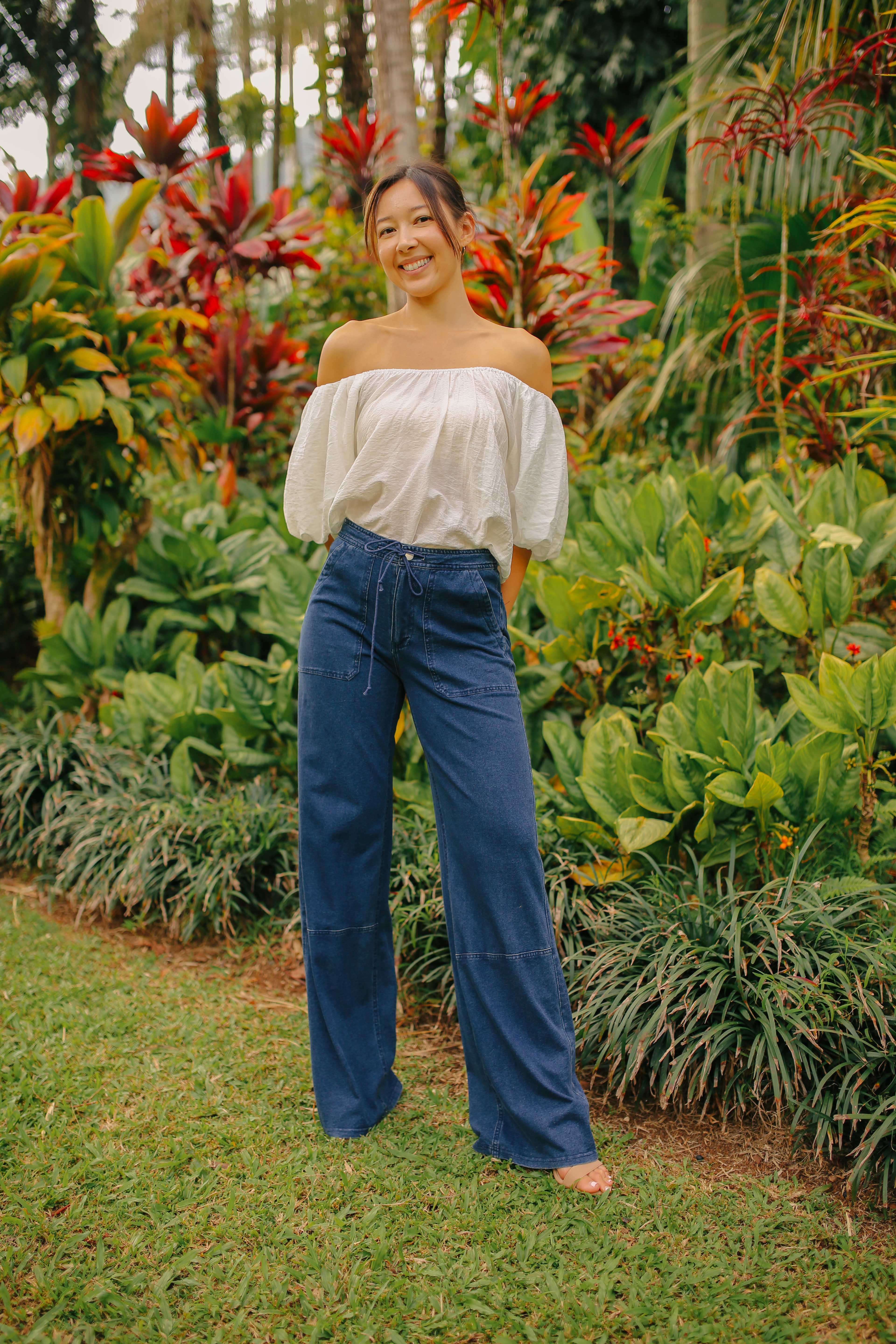 A woman standing outdoors wearing blue wide-leg cargo pants and a white off-shoulder top.