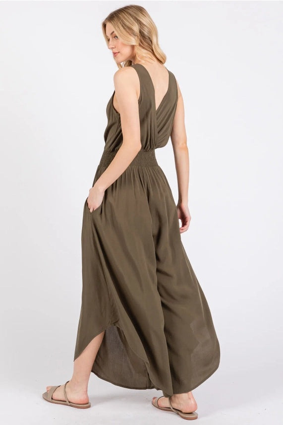 sleeveless, olive green jumpsuit with a smocked waist and wide, flowing legs. She is standing with one hand in a pocket and is facing sideways. She is also wearing a pair of sandals.