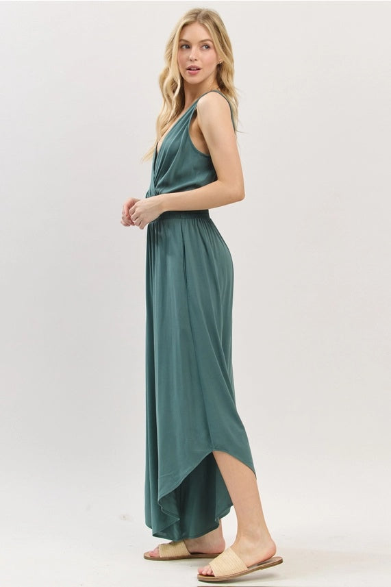 A side view of a young woman modeling a sleeveless, sea-green jumpsuit with a smocked waist and wide, flowing legs with a high-low hem.