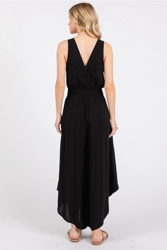 A back view of a woman wearing a black, sleeveless jumpsuit with a V-neckline and a smocked waistband. The jumpsuit features wide, flowy legs with a high-low hemline.