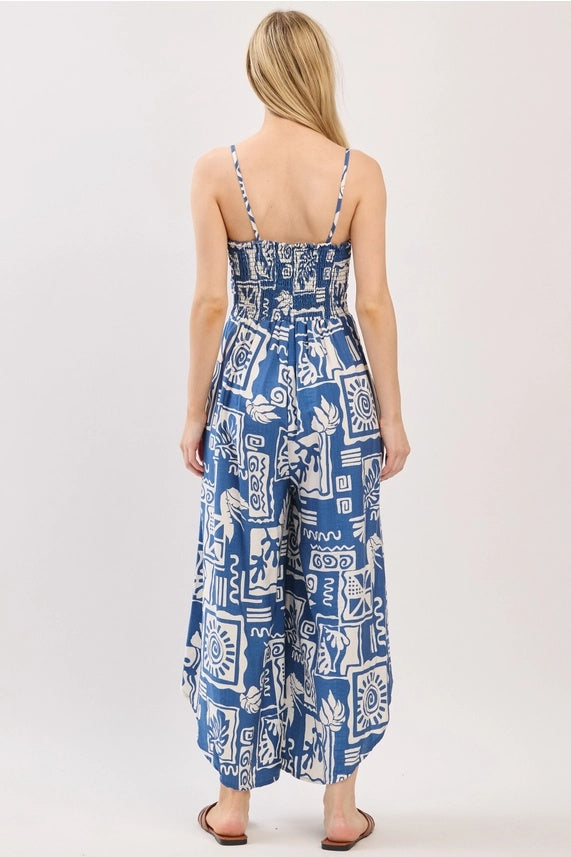 
A rear view of a young woman modeling a blue and white patterned sleeveless jumpsuit with a smocked back and wide legs. The pattern is abstract and geometric.