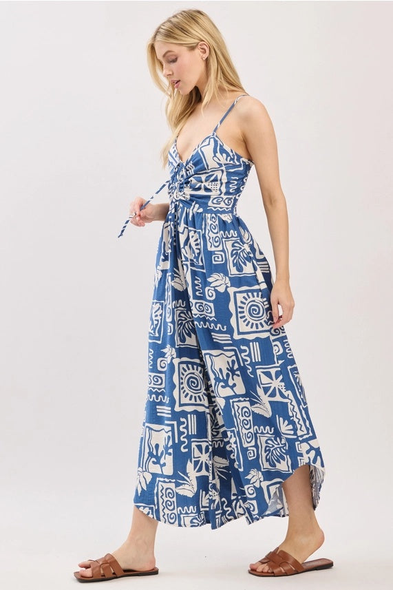 
A side view of a young woman modeling a blue and white patterned sleeveless jumpsuit with a V-neck, cinched waist, and wide legs. She is holding one of the drawstrings on the bust.